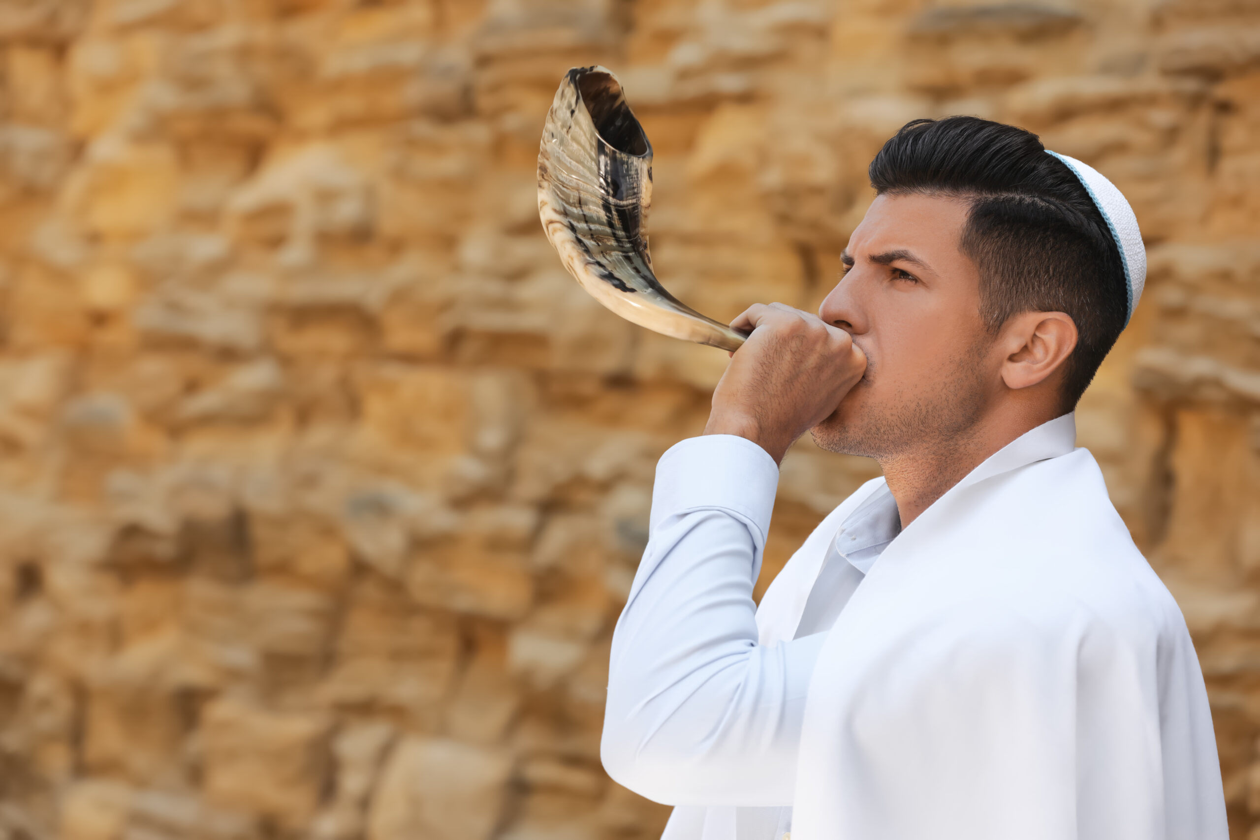 Jewish man in kippah and tallit blowing shofar outdoors. Rosh Hashanah celebration